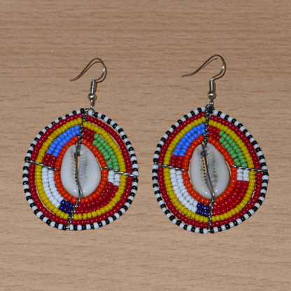 Boucles d'oreilles africaines ethniques composées de perles multicolores et de cauris montés en forme de boucliers Massai.  Elles mesurent 7 cm de long et 4 cm de large et se portent sur des oreilles percées.