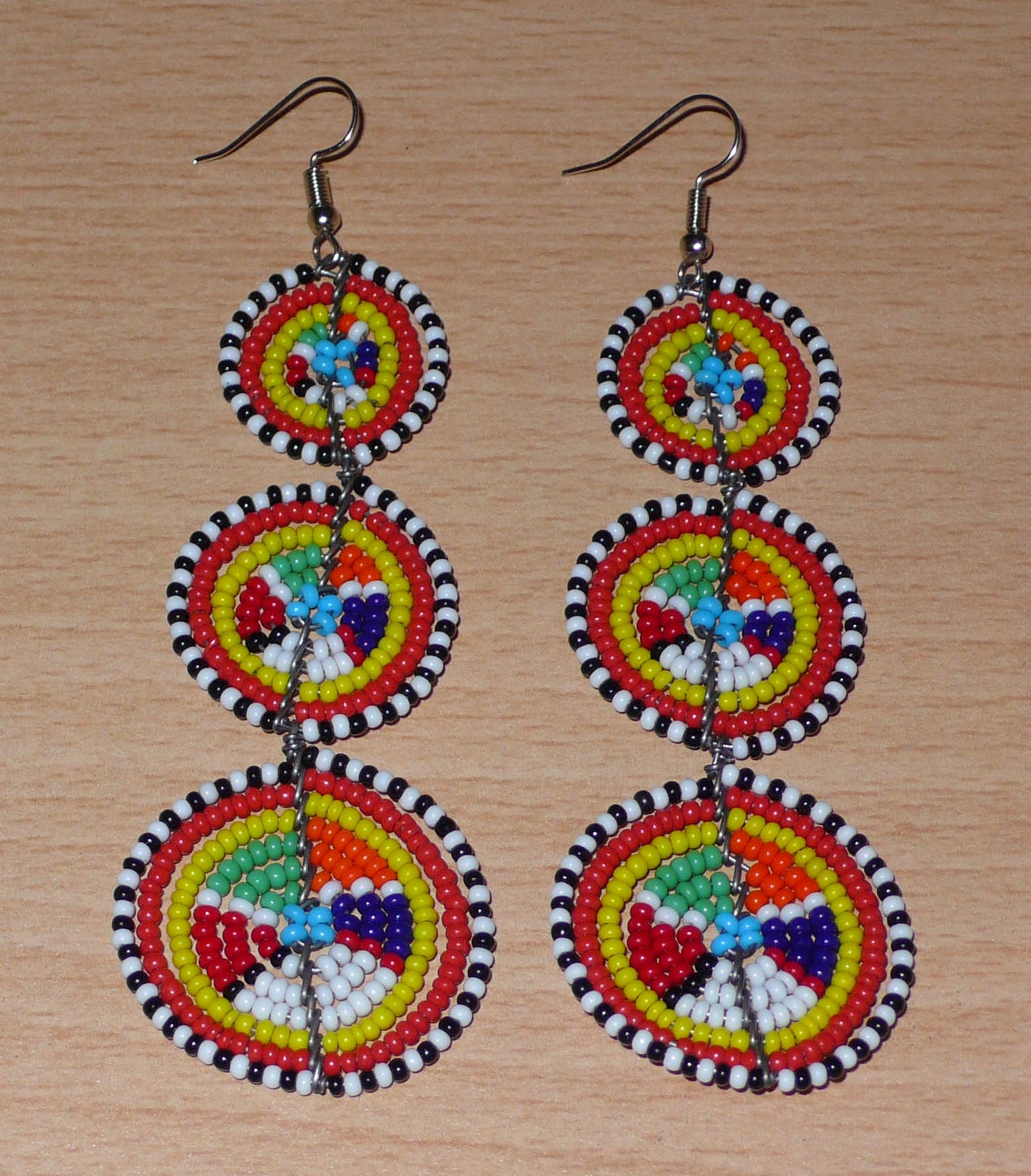 Ajoutez une touche colorée à votre look, évocatrice du style afro chic des années 70, avec ces boucles d'oreilles africaines traditionnelles de confection artisanale Massai. Elles se composent de perles de rocaille multicolores glissées sur des fils de fer montés en forme de trois cercles superposés. Ces boucles d'oreilles mesurent 10.5 cm de long et 3 cm de large ; elles se portent avec des crochets en acier inoxydable sur des oreilles percées.