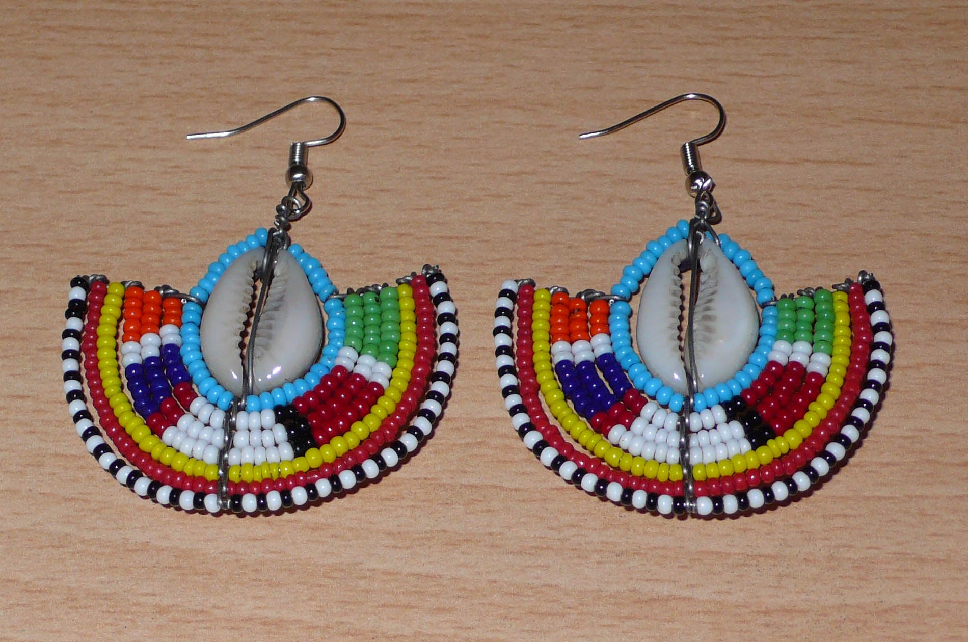 Affirmez votre style et apportez une touche exotique à votre look avec cette paire de boucles d'oreilles africaines Massai issues de l'artisanat traditionnel kenyan. Ces boucles d'oreilles aux accents ethniques chic se composent de perles de rocaille bleues et multicolores et de coquillages cauris. De taille moyenne, elles mesurent 6 cm de long et 4.5 cm de large, et se portent avec des crochets en acier inoxydable sur des oreilles percées.