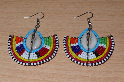 Affirmez votre style et apportez une touche exotique à votre look avec cette paire de boucles d'oreilles africaines Massai issues de l'artisanat traditionnel kenyan. Ces boucles d'oreilles aux accents ethniques chic se composent de perles de rocaille bleues et multicolores et de coquillages cauris. De taille moyenne, elles mesurent 6 cm de long et 4.5 cm de large, et se portent avec des crochets en acier inoxydable sur des oreilles percées.