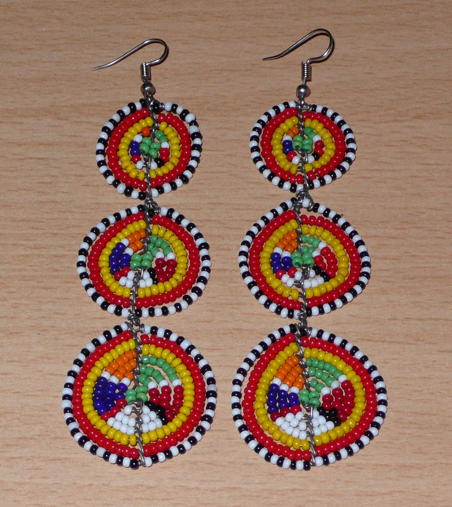 Affirmez votre style avec ces boucles d'oreilles africaines traditionnelles de confection artisanale Massai, évoquant le style afro chic des années 70. Elles se composent de perles de rocaille multicolores enfilées sur des fils de fer montés en forme de trois cercles superposés. Ces boucles d'oreilles mesurent 11 cm de long et 3.5 cm de large ; elles se portent avec des crochets en acier inoxydable sur des oreilles percées.