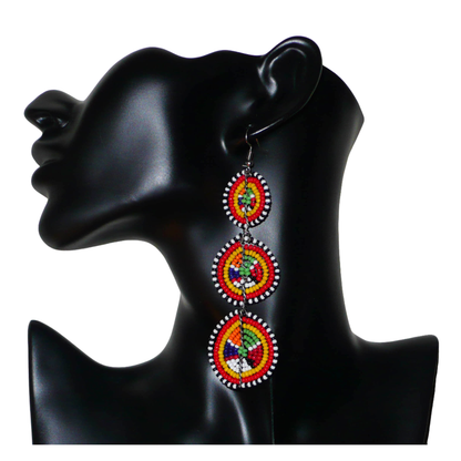 Affirmez votre style avec ces boucles d'oreilles africaines traditionnelles de confection artisanale Massai, évoquant le style afro chic des années 70. Elles se composent de perles de rocaille multicolores enfilées sur des fils de fer montés en forme de trois cercles superposés. Ces boucles d'oreilles mesurent 11 cm de long et 3.5 cm de large ; elles se portent avec des crochets en acier inoxydable sur des oreilles percées.