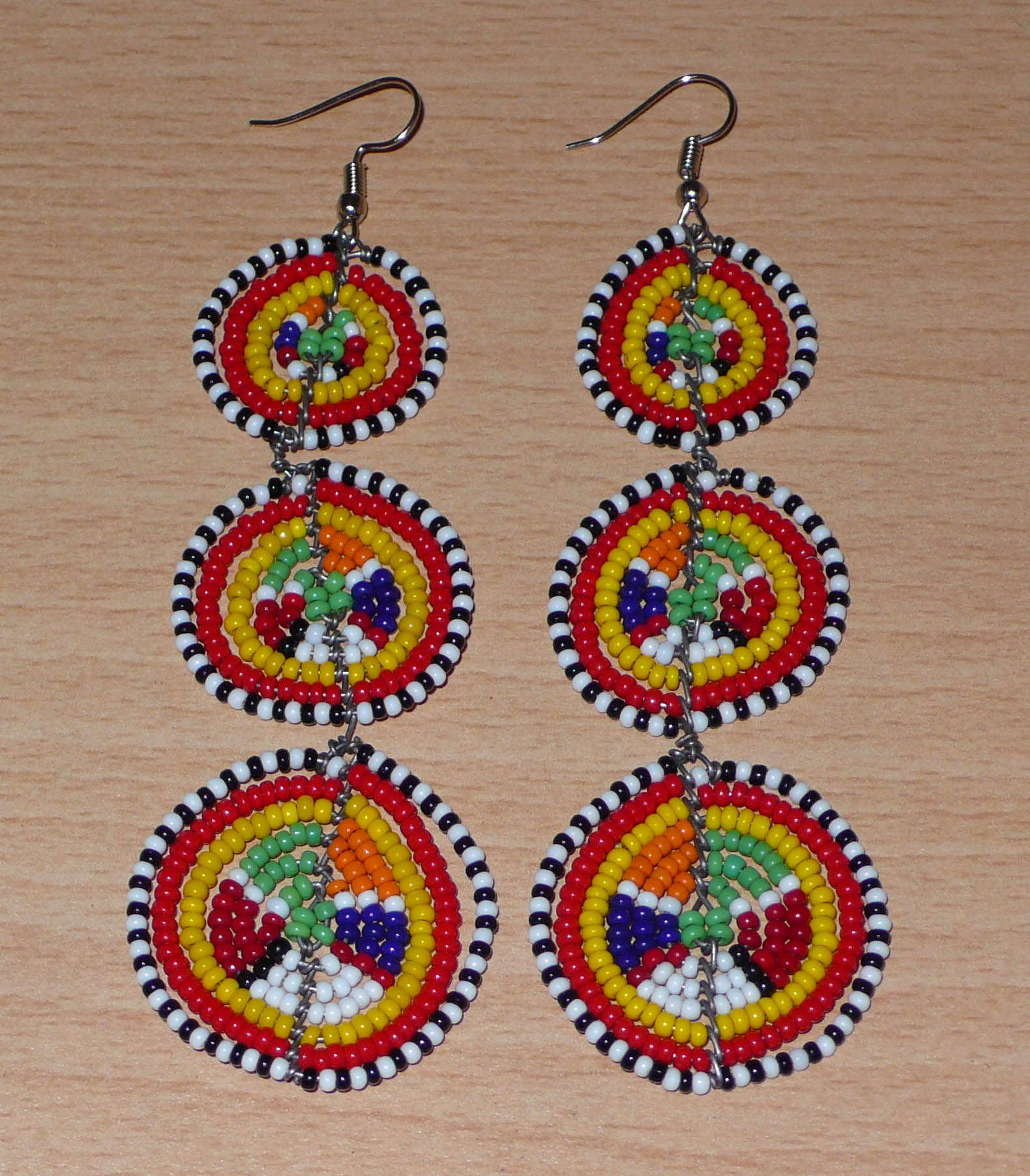 Affirmez votre style avec ces boucles d'oreilles africaines traditionnelles de confection artisanale Massai, évoquant le style afro chic des années 70. Elles se composent de perles de rocaille multicolores enfilées sur des fils de fer montés en forme de trois cercles superposés. Ces boucles d'oreilles mesurent 11 cm de long et 3.5 cm de large ; elles se portent avec des crochets en acier inoxydable sur des oreilles percées.