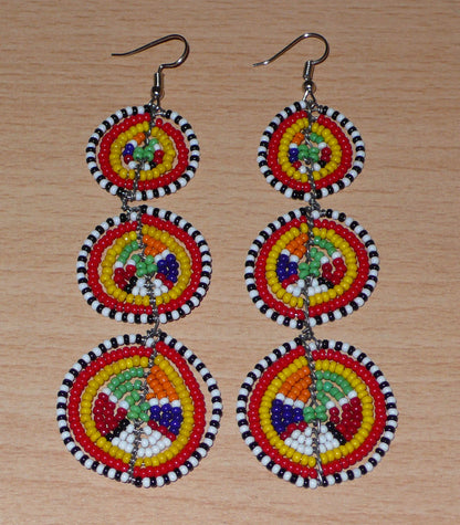 Affirmez votre style avec ces boucles d'oreilles africaines traditionnelles de confection artisanale Massai, évoquant le style afro chic des années 70. Elles se composent de perles de rocaille multicolores enfilées sur des fils de fer montés en forme de trois cercles superposés. Ces boucles d'oreilles mesurent 11 cm de long et 3.5 cm de large ; elles se portent avec des crochets en acier inoxydable sur des oreilles percées.