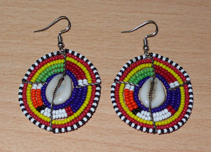 Boucles d'oreilles africaines traditionnelles aux motifs ethniques caractéristiques de l'artisanat Massai. De fabrication kenyane, elles se composent de perles de rocaille bleu foncé et multicolores et de cauris fixés sur des fils de fer montés en forme de boucliers Massai. Ces boucles d'oreilles de taille moyenne mesurent 6 cm de long et 4 cm de large ; elles se portent avec des crochets en acier inoxydable sur des oreilles percées.