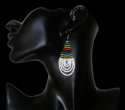 Boucles d'oreilles africaines à motifs traditionnels zoulous en forme de gouttes composées d'un tissage de perles de rocaille blanches et multicolores. Elles mesurent 7 cm de long et 2,5 cm de large et se portent sur des oreilles percées.