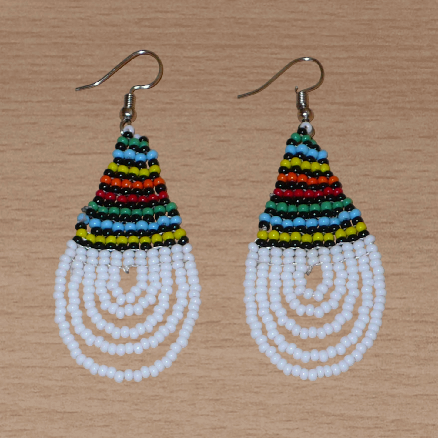 Boucles d'oreilles africaines à motifs traditionnels zoulous en forme de gouttes composées d'un tissage de perles de rocaille blanches et multicolores. Elles mesurent 7 cm de long et 2,5 cm de large et se portent sur des oreilles percées.