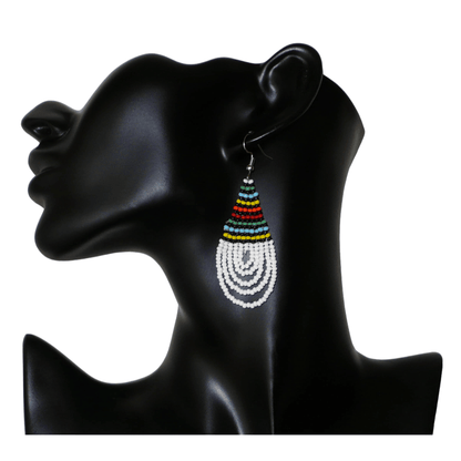 Boucles d'oreilles africaines à motifs traditionnels zoulous en forme de gouttes composées d'un tissage de perles de rocaille blanches et multicolores. Elles mesurent 7 cm de long et 2,5 cm de large et se portent sur des oreilles percées.