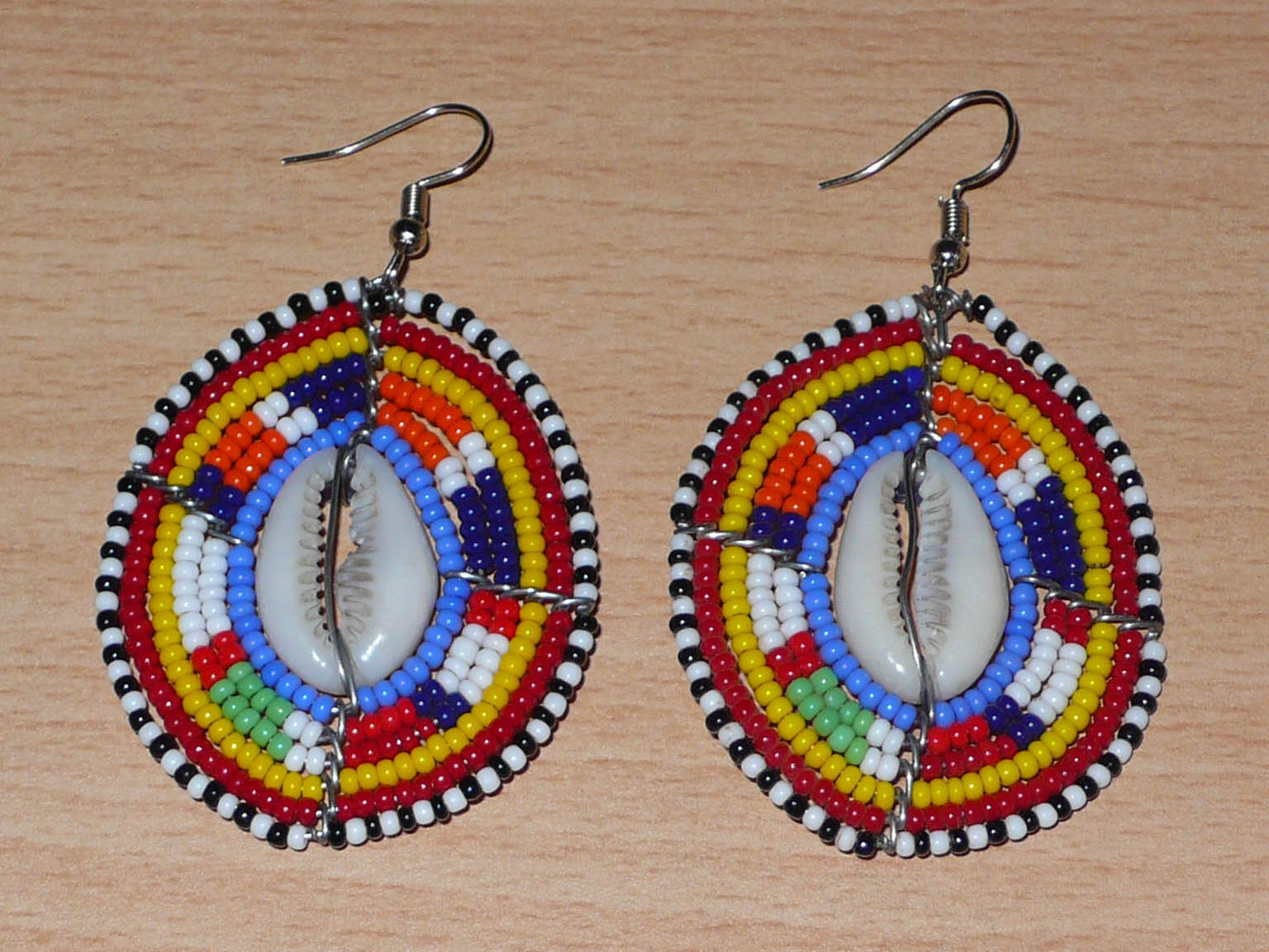 Boucles d'oreilles africaines en forme de boucliers Massai composées de perles de rocaille multicolores et de coquillages cauris. Elles mesurent 7 cm de long et 4 cm de large.