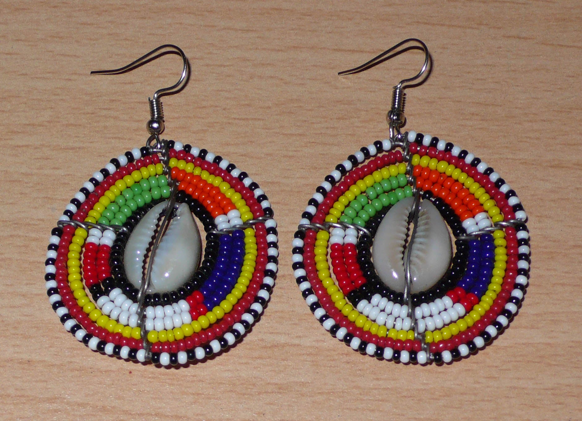 Boucles d'oreilles africaines Massai à motifs ethniques traditionnels : de fabrication artisanale kenyane, elles se composent de perles de rocaille noires et multicolores et de coquillages cauris fixés sur des fils de fer montés en forme de boucliers Massai. De taille moyenne, ces boucles d'oreilles aux accents exotiques mesurent 6.5 cm de long et 4 cm de large ; elles se portent avec des crochets en acier inoxydable sur des oreilles percées.