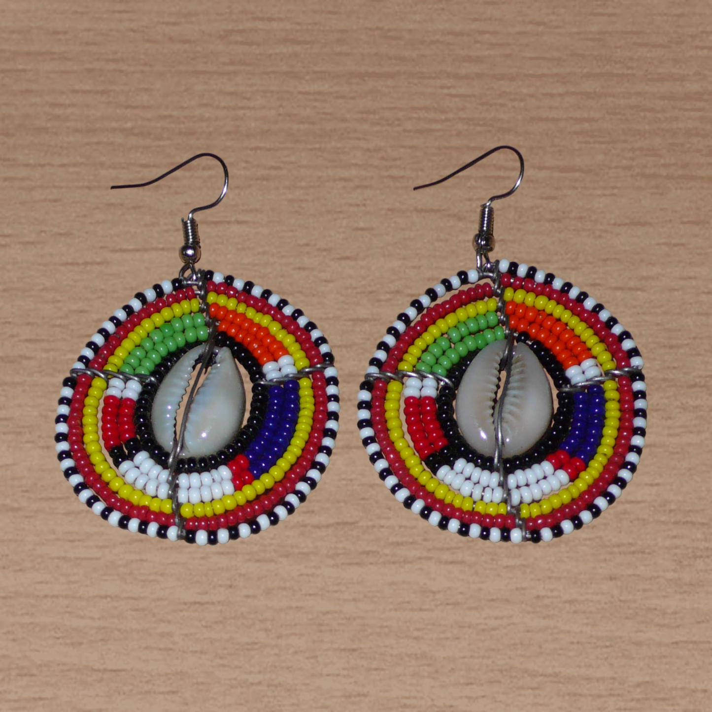 Boucles d'oreilles africaines Massai à motifs ethniques traditionnels : de fabrication artisanale kenyane, elles se composent de perles de rocaille noires et multicolores et de coquillages cauris fixés sur des fils de fer montés en forme de boucliers Massai. De taille moyenne, ces boucles d'oreilles aux accents exotiques mesurent 6.5 cm de long et 4 cm de large ; elles se portent avec des crochets en acier inoxydable sur des oreilles percées.