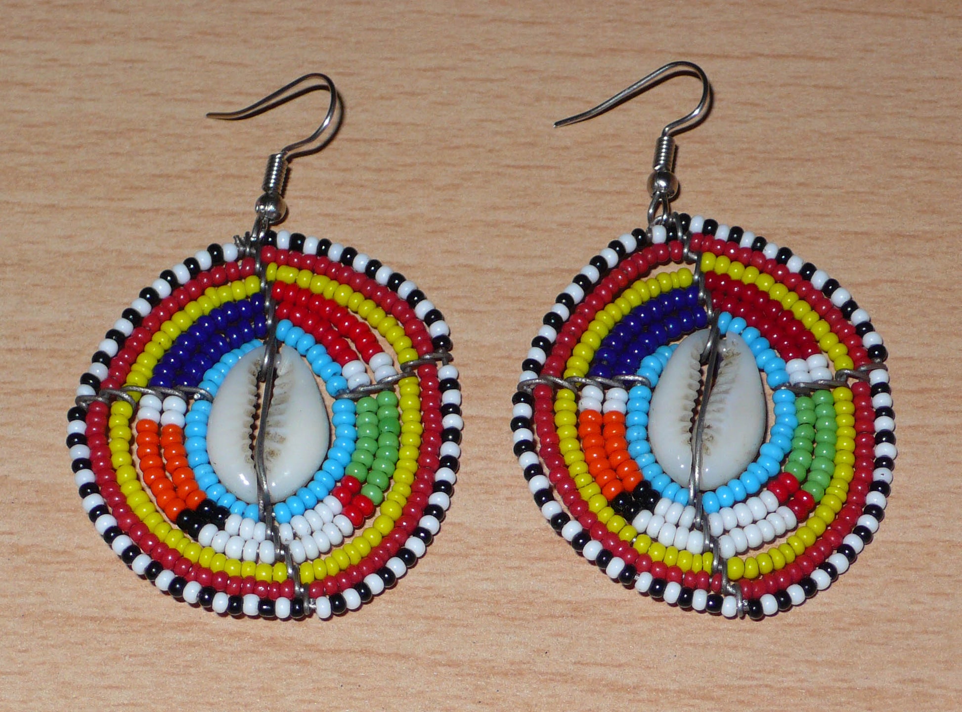 Boucles d'oreilles africaines traditionnelles aux motifs ethniques caractéristiques de l'artisanat Massai. De fabrication kenyane, elles se composent de perles de rocaille bleues et multicolores et de coquillages fixés sur des fils de fer montés en forme de boucliers Massai. Ces boucles d'oreilles de taille moyenne mesurent 6.5 cm de long et 4 cm de large ; elles se portent avec des crochets en acier inoxydable sur des oreilles percées.