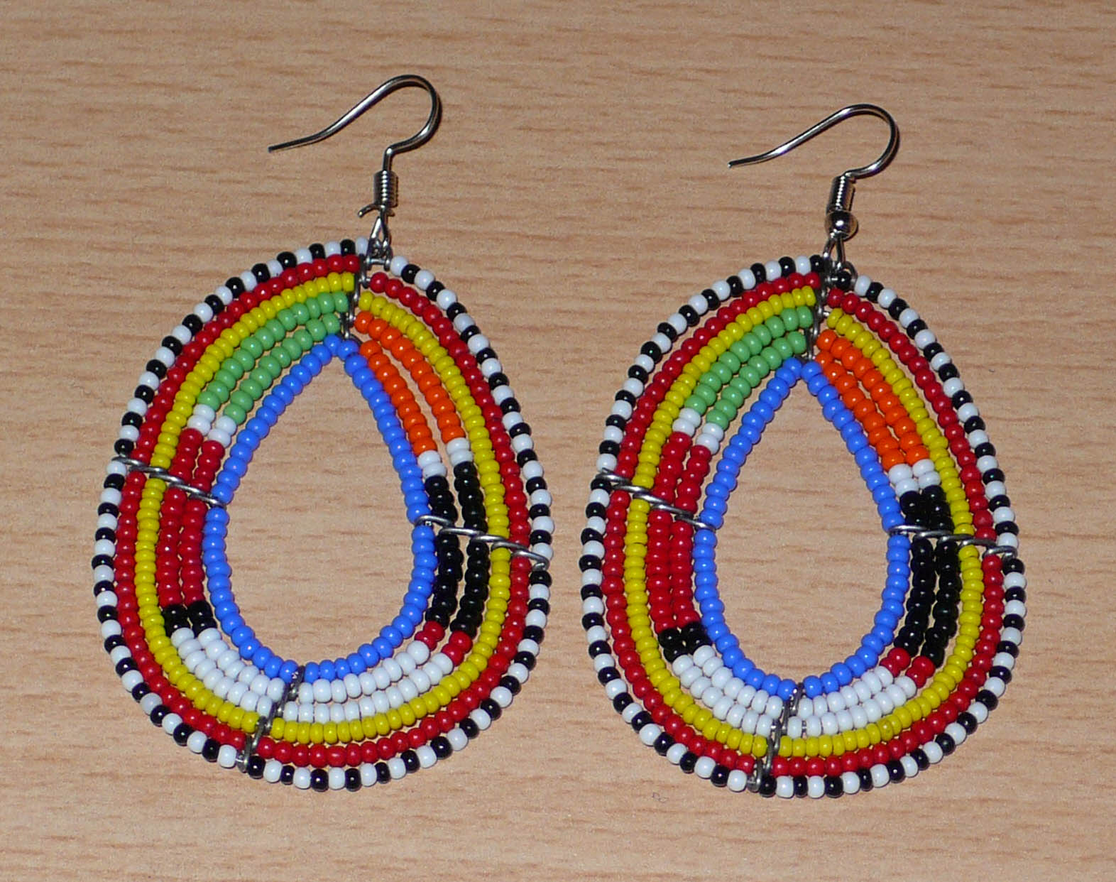 Boucles d'oreilles ethniques multi-rangs à motifs traditionnels Massai composées de perles de rocaille bleues et multicolores glissées sur six rangs de fils de fer montés en forme de gouttes. Elles mesurent 7.5 cm de long et 4 cm de large, et se portent avec des crochets en acier inoxydable sur des oreilles percées.