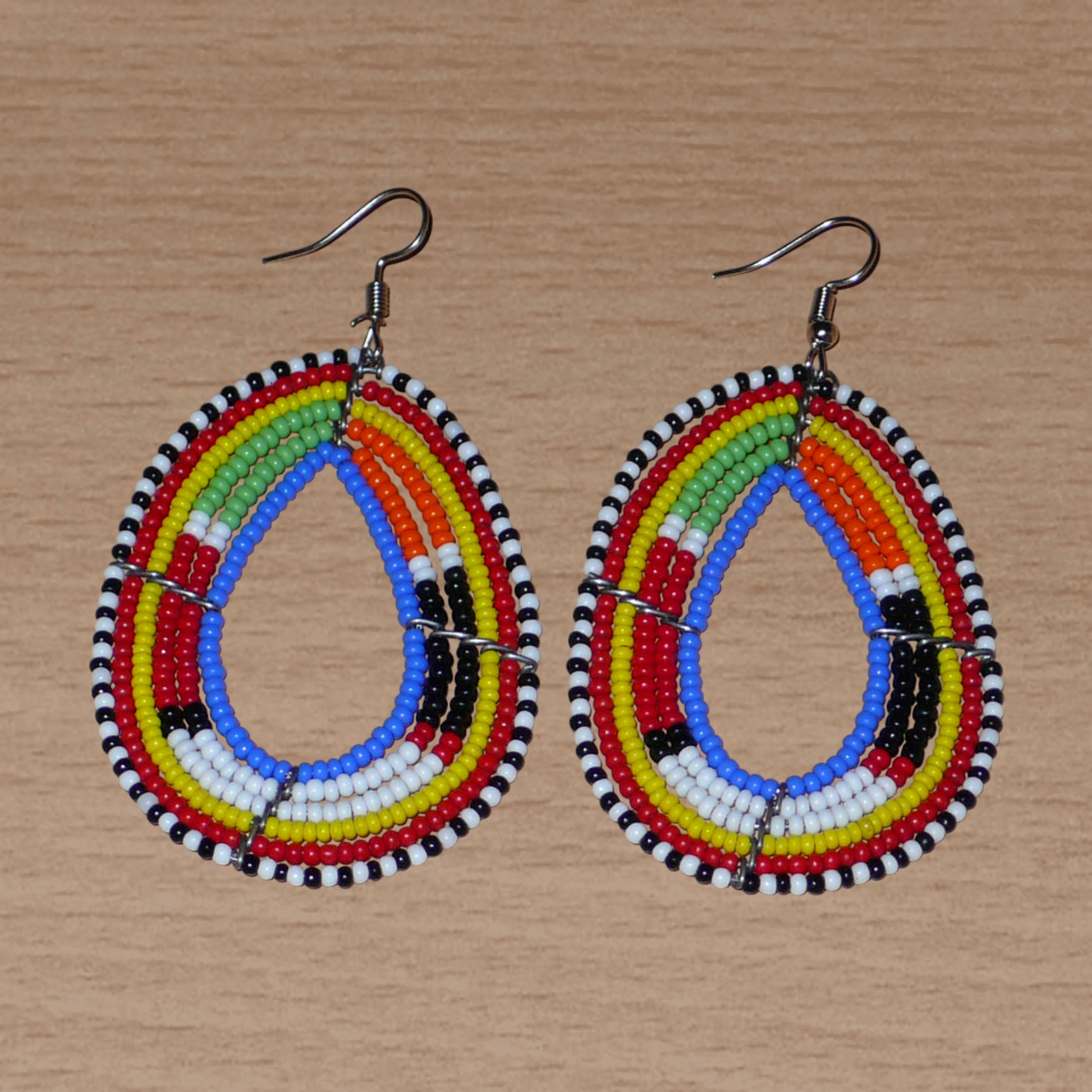 Boucles d'oreilles ethniques multi-rangs à motifs traditionnels Massai composées de perles de rocaille bleues et multicolores glissées sur six rangs de fils de fer montés en forme de gouttes. Elles mesurent 7.5 cm de long et 4 cm de large, et se portent avec des crochets en acier inoxydable sur des oreilles percées.