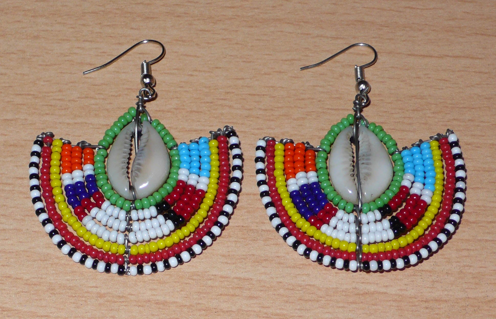 Ajoutez une touche afro chic à votre style avec cette jolie paire de boucles d'oreilles africaines issues de l'artisanat traditionnel Massai. Ces boucles d'oreilles aux accents exotiques se composent de perles de rocaille vertes et multicolores et de coquillages cauris. De taille moyenne, elles mesurent 6 cm de long et 4.5 cm de large, et se portent avec des crochets en acier inoxydable sur des oreilles percées.