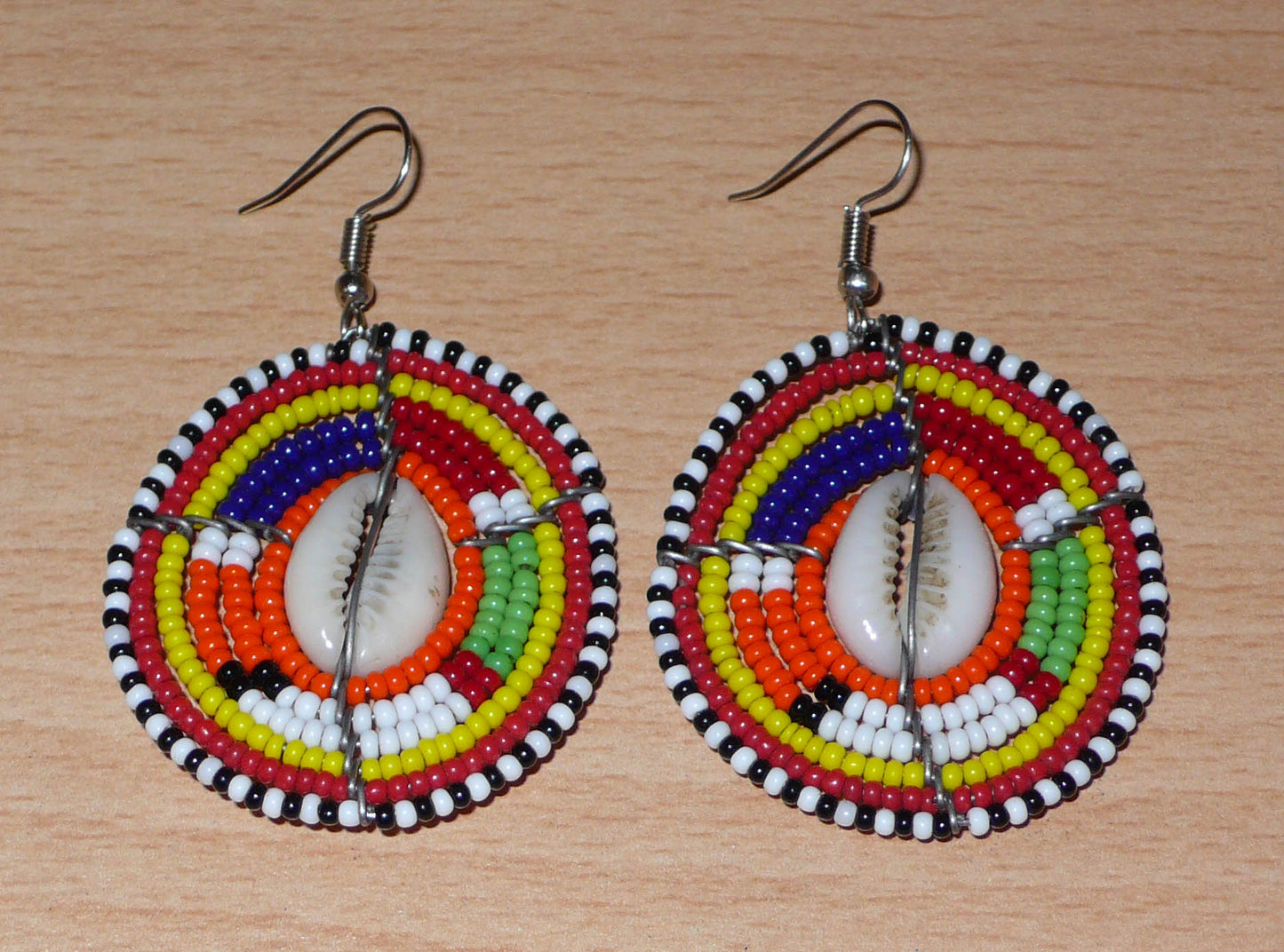Boucles d'oreilles africaines aux couleurs exotiques et motifs ethniques caractéristiques de l'artisanat traditionnel Massai. Confectionnées au Kenya, elles se composent de perles de rocaille orange et multicolores et de coquillages fixés sur des fils de fer montés en forme de boucliers Massai. Ces boucles d'oreilles de taille moyenne mesurent 6 cm de long et 4 cm de large, et se portent avec des crochets en acier inoxydable sur des oreilles percées.