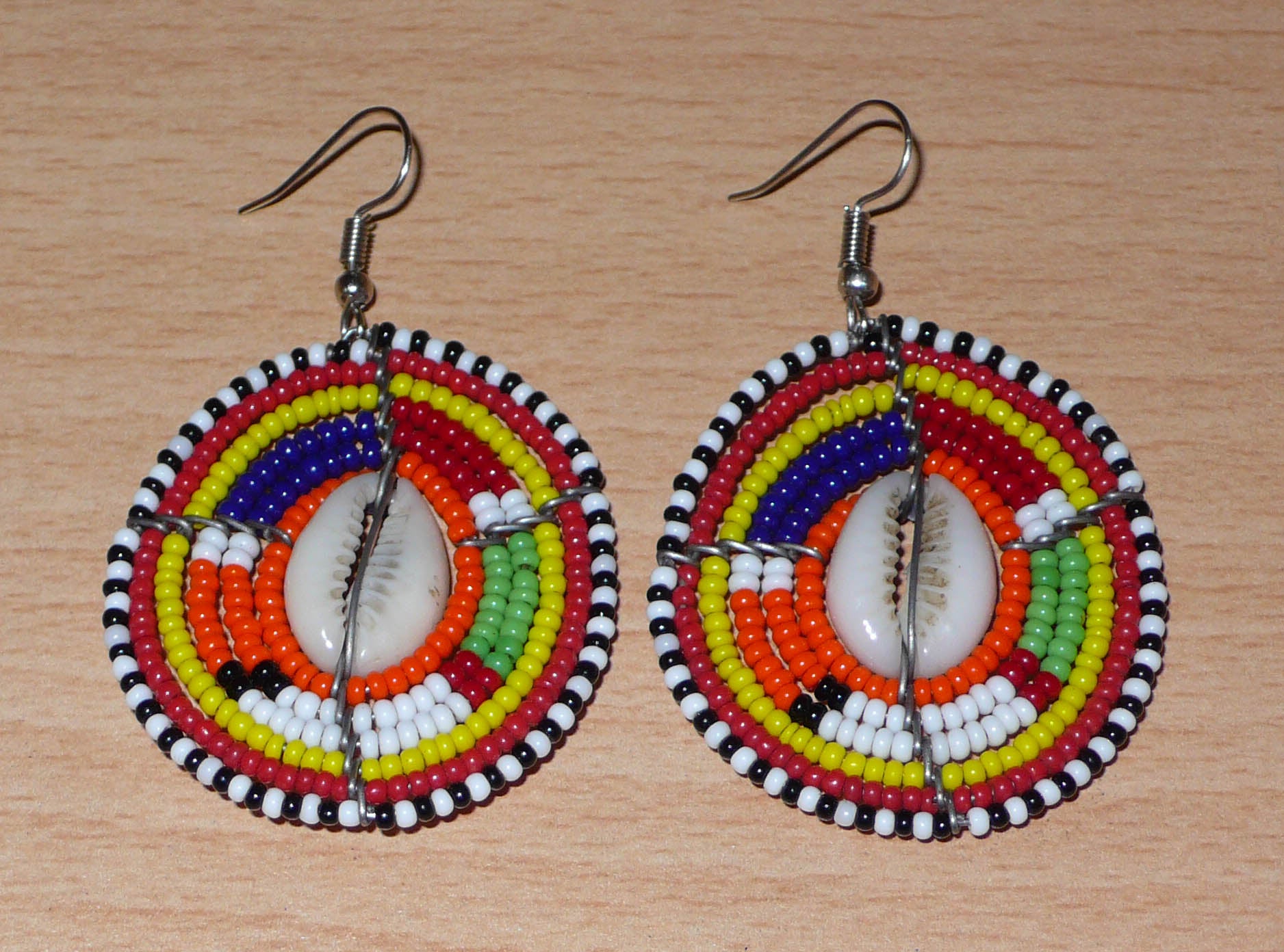 Boucles d'oreilles africaines aux couleurs exotiques et motifs ethniques caractéristiques de l'artisanat traditionnel Massai. Confectionnées au Kenya, elles se composent de perles de rocaille orange et multicolores et de coquillages fixés sur des fils de fer montés en forme de boucliers Massai. Ces boucles d'oreilles de taille moyenne mesurent 6 cm de long et 4 cm de large, et se portent avec des crochets en acier inoxydable sur des oreilles percées.