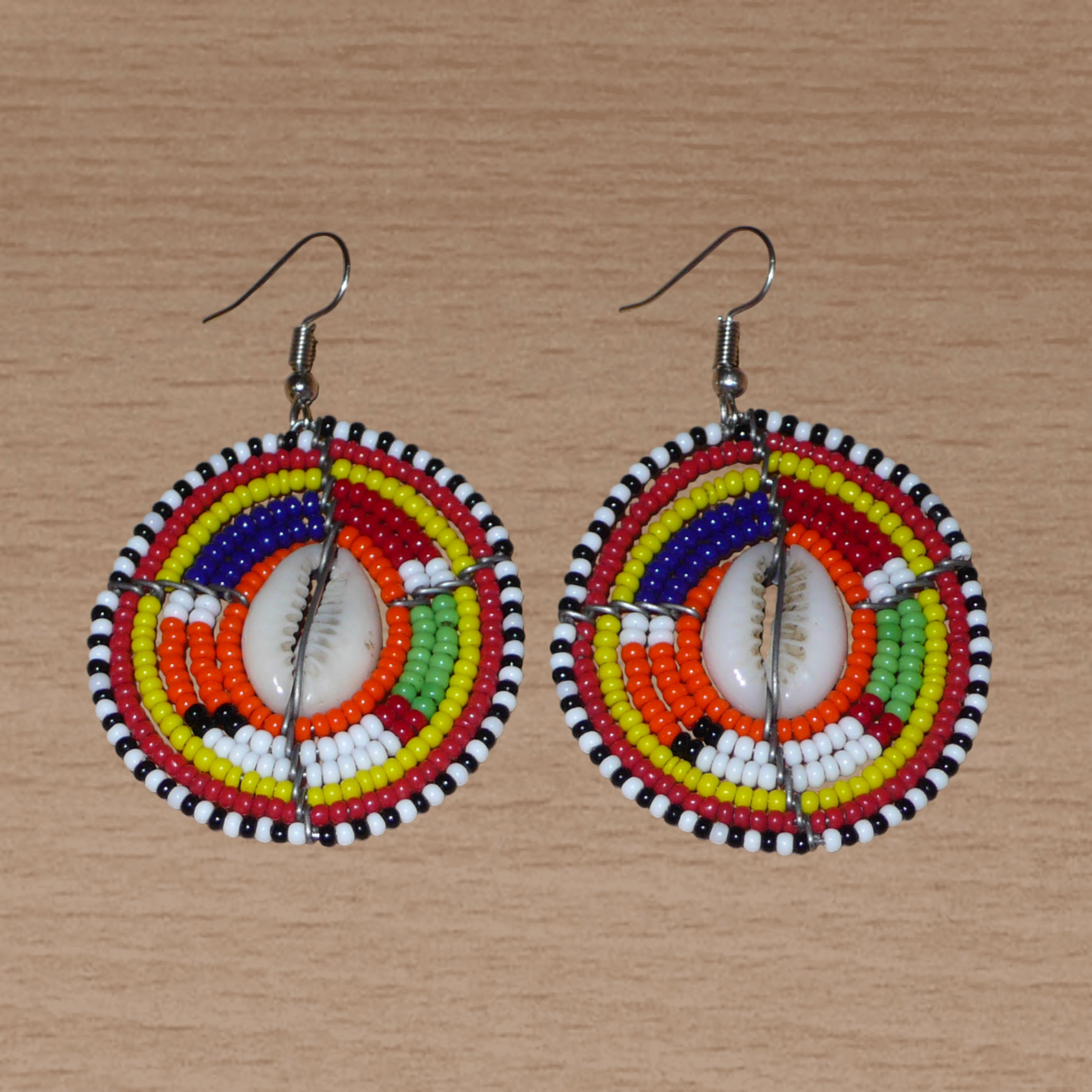 Boucles d'oreilles africaines aux couleurs exotiques et motifs ethniques caractéristiques de l'artisanat traditionnel Massai. Confectionnées au Kenya, elles se composent de perles de rocaille orange et multicolores et de coquillages fixés sur des fils de fer montés en forme de boucliers Massai. Ces boucles d'oreilles de taille moyenne mesurent 6 cm de long et 4 cm de large, et se portent avec des crochets en acier inoxydable sur des oreilles percées.