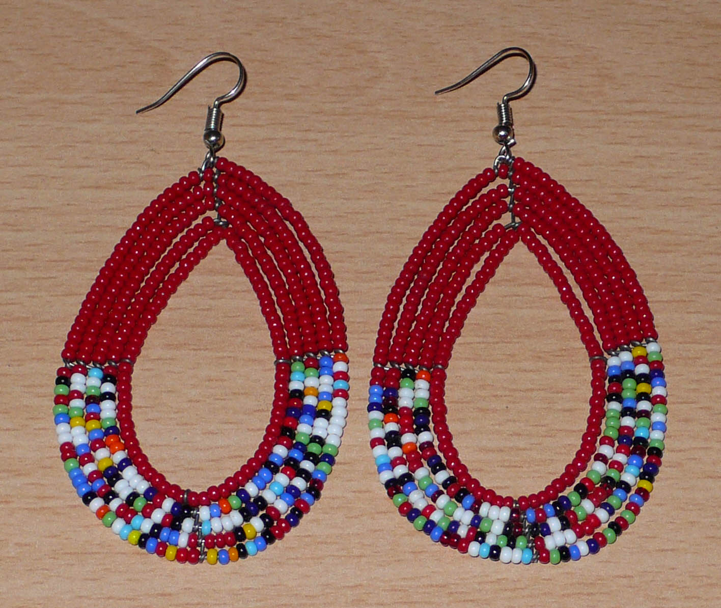 Longues boucles d'oreilles rouges et multicolores en forme de gouttes composées de perles de rocaille. Elles mesurent 9 cm de long et 4,5 cm de large et se portent avec des crochets sur des oreilles percées.