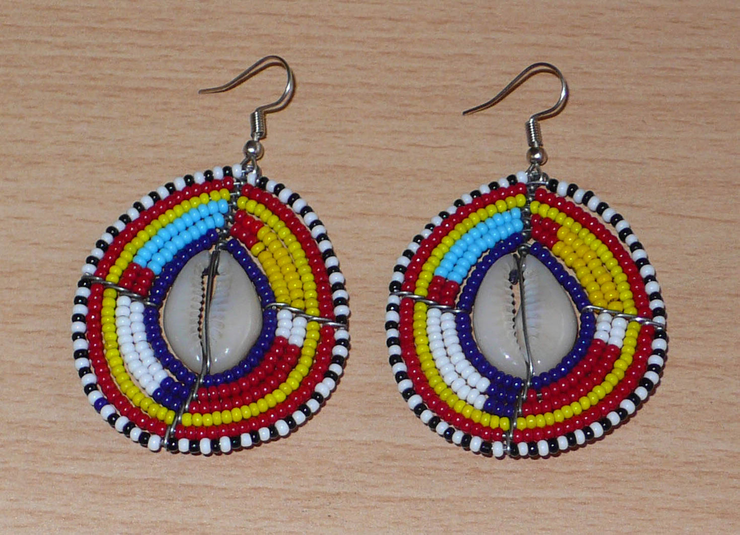 Boucles d'oreilles africaines traditionnelles aux couleurs et motifs ethniques Massai. Elles se composent de perles de rocaille multicolores enfilées sur des fils de fer montés en forme de boucliers Massai ; des coquillages sont incrustés en leur centre. Elles mesurent 7 cm de long et 4 cm de large et se portent avec des crochets en acier inoxydable sur des oreilles percées.
