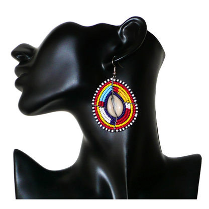 Boucles d'oreilles africaines traditionnelles aux couleurs et motifs ethniques Massai. Elles se composent de perles de rocaille multicolores enfilées sur des fils de fer montés en forme de boucliers Massai ; des coquillages sont incrustés en leur centre. Elles mesurent 7 cm de long et 4 cm de large et se portent avec des crochets en acier inoxydable sur des oreilles percées.