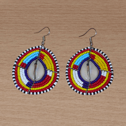 Boucles d'oreilles africaines traditionnelles aux couleurs et motifs ethniques Massai. Elles se composent de perles de rocaille multicolores enfilées sur des fils de fer montés en forme de boucliers Massai ; des coquillages sont incrustés en leur centre. Elles mesurent 7 cm de long et 4 cm de large et se portent avec des crochets en acier inoxydable sur des oreilles percées.