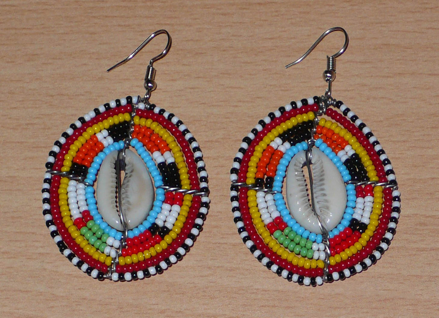 Boucles d'oreilles africaines ethniques en perles de rocaille colorées et coquillages cauris montés en forme de boucliers Massai. Elles mesurent 7 cm de long et 4.5 cm de large, et se portent avec des crochets en acier inoxydable sur des oreilles percées. 