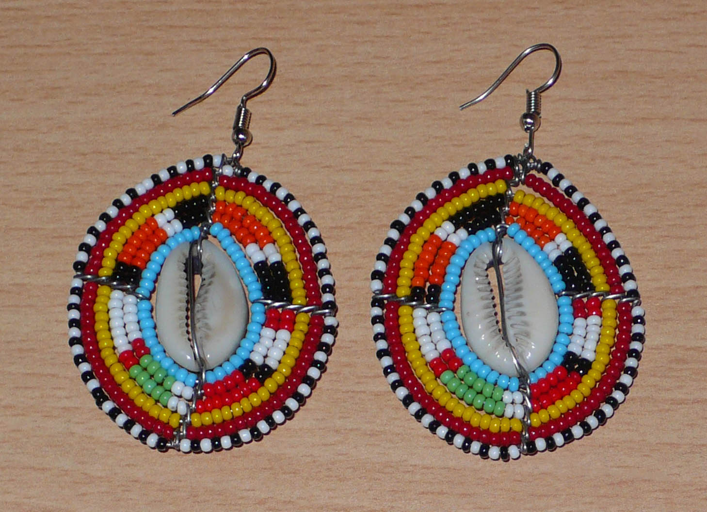 Boucles d'oreilles africaines ethniques en perles de rocaille colorées et coquillages cauris montés en forme de boucliers Massai. Elles mesurent 7 cm de long et 4.5 cm de large, et se portent avec des crochets en acier inoxydable sur des oreilles percées. 