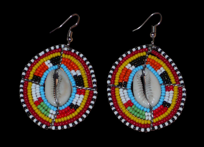 Boucles d'oreilles africaines ethniques en perles de rocaille colorées et coquillages cauris montés en forme de boucliers Massai. Elles mesurent 7 cm de long et 4.5 cm de large, et se portent avec des crochets en acier inoxydable sur des oreilles percées. 
