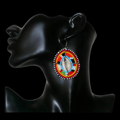Boucles d'oreilles africaines ethniques en perles de rocaille colorées et coquillages cauris montés en forme de boucliers Massai. Elles mesurent 7 cm de long et 4.5 cm de large, et se portent avec des crochets en acier inoxydable sur des oreilles percées. 