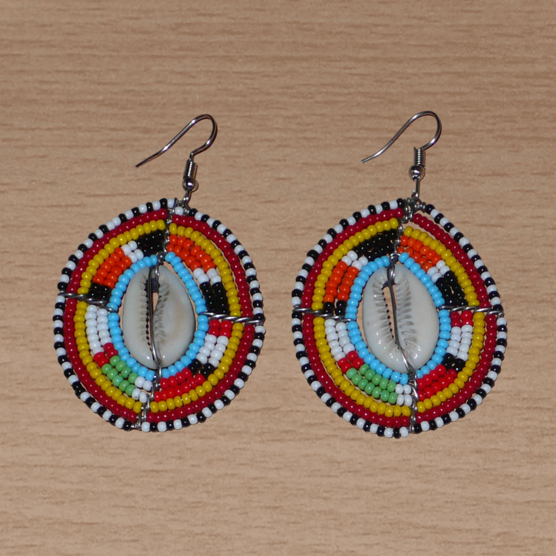 Boucles d'oreilles africaines ethniques en perles de rocaille colorées et coquillages cauris montés en forme de boucliers Massai. Elles mesurent 7 cm de long et 4.5 cm de large, et se portent avec des crochets en acier inoxydable sur des oreilles percées. 