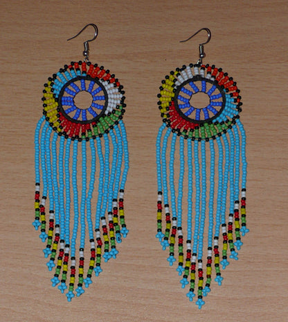 Longues boucles d'oreilles sud-africaines traditionnelles à franges et motifs zoulous composées de perles de rocaille bleu ciel et multicolores. Elles mesurent 14,5 cm de long et 4,5 cm de large, et se portent avec des crochets en acier inoxydable sur des oreilles percées.