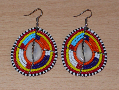 Boucles d'oreilles africaines Massai composées de perles de rocaille multicolores et de coquillages cauris. Elles mesurent 7 cm de long et 4 cm de large et se portent avec des crochets en acier inoxydable sur des oreilles percées.