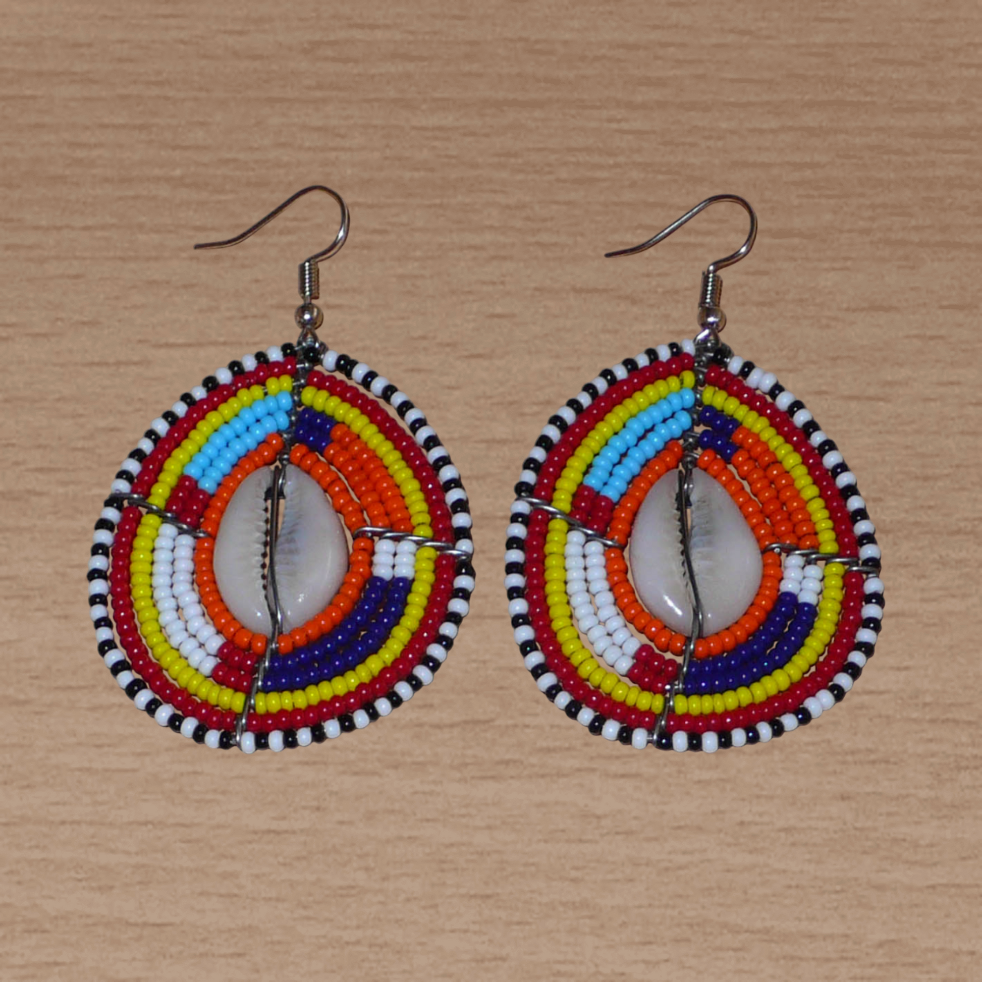 Boucles d'oreilles africaines Massai composées de perles de rocaille multicolores et de coquillages cauris. Elles mesurent 7 cm de long et 4 cm de large et se portent avec des crochets en acier inoxydable sur des oreilles percées.