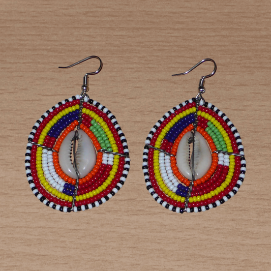 Boucles d'oreilles africaines à motifs ethniques composées de perles et de coquillages montés en forme de boucliers Massai. Elles mesurent 7 cm de long et 4 cm de large et se portent sur des oreilles percées. 