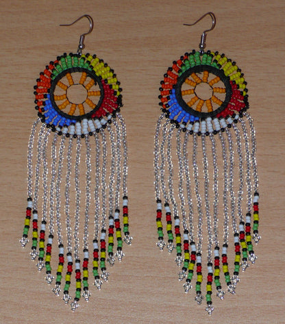 Longues boucles d'oreilles sud-africaines traditionnelles à franges et motifs zoulous composées de perles de rocaille gris clair translucides et multicolores. Elles mesurent 14,5 cm de long et 4,5 cm de large, et elles se portent avec des crochets en acier inoxydable sur des oreilles percées.