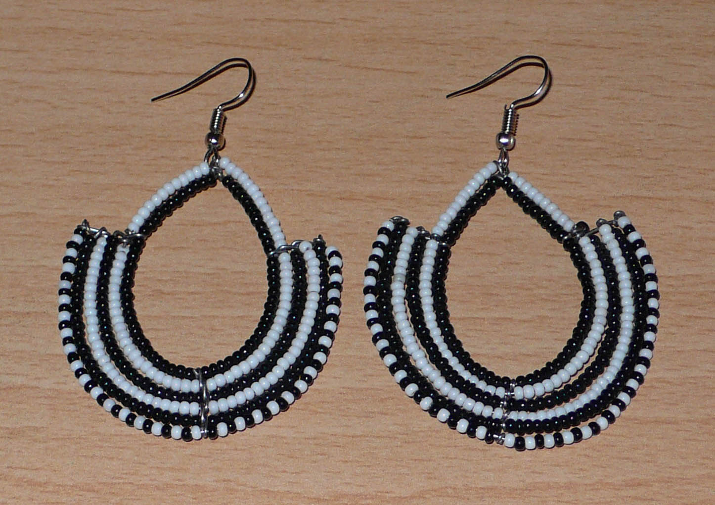 Boucles d'oreilles ethniques bicolores composées de perles de rocaille noires et blanches glissées sur des structures en fils de fer montés en forme d'ancres. Elles mesurent 7 cm de long et 4,5 cm de large et se portent sur des oreilles percées.