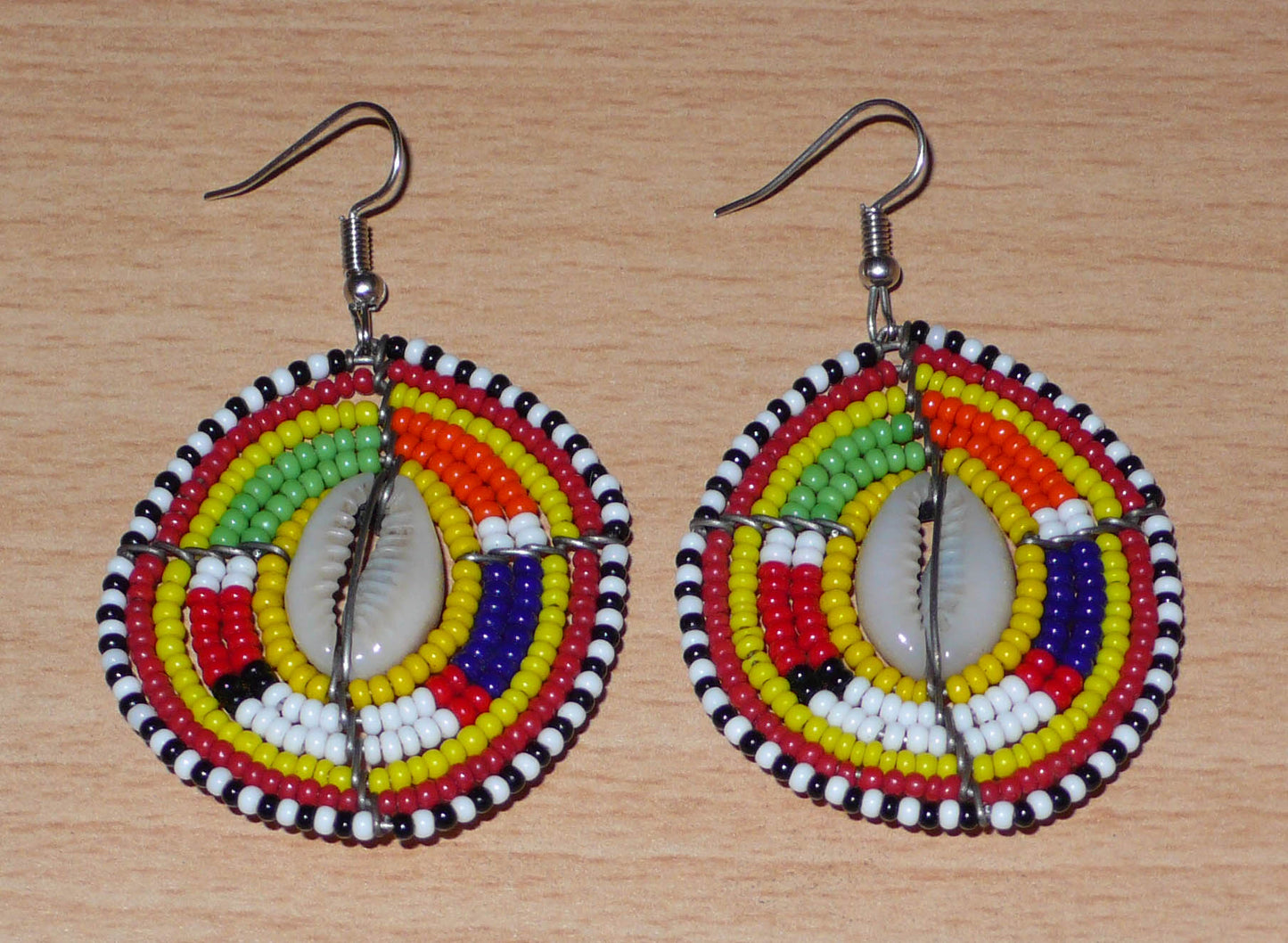 Ambiance exotique avec ces jolies boucles d'oreilles africaines à motifs ethniques traditionnels Massai de conception artisanale kenyane. Elles sont composées de perles de rocaille jaunes et multicolores et de coquillages cauris fixés sur des fils de fer montés en forme de boucliers Massai. Ces boucles d'oreilles de taille moyenne mesurent 6.5 cm de long et 4 cm de large ; elles se portent avec des crochets en acier inoxydable sur des oreilles percées.