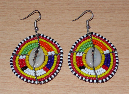 Ambiance exotique avec ces jolies boucles d'oreilles africaines à motifs ethniques traditionnels Massai de conception artisanale kenyane. Elles sont composées de perles de rocaille jaunes et multicolores et de coquillages cauris fixés sur des fils de fer montés en forme de boucliers Massai. Ces boucles d'oreilles de taille moyenne mesurent 6.5 cm de long et 4 cm de large ; elles se portent avec des crochets en acier inoxydable sur des oreilles percées.