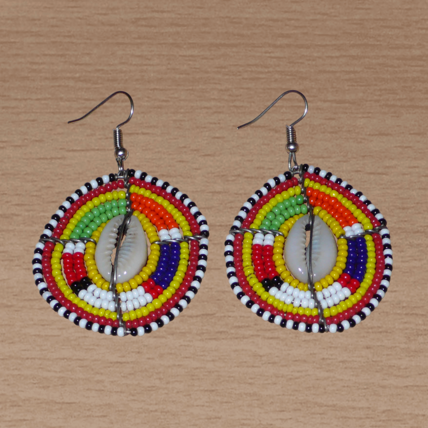 Ambiance exotique avec ces jolies boucles d'oreilles africaines à motifs ethniques traditionnels Massai de conception artisanale kenyane. Elles sont composées de perles de rocaille jaunes et multicolores et de coquillages cauris fixés sur des fils de fer montés en forme de boucliers Massai. Ces boucles d'oreilles de taille moyenne mesurent 6.5 cm de long et 4 cm de large ; elles se portent avec des crochets en acier inoxydable sur des oreilles percées.