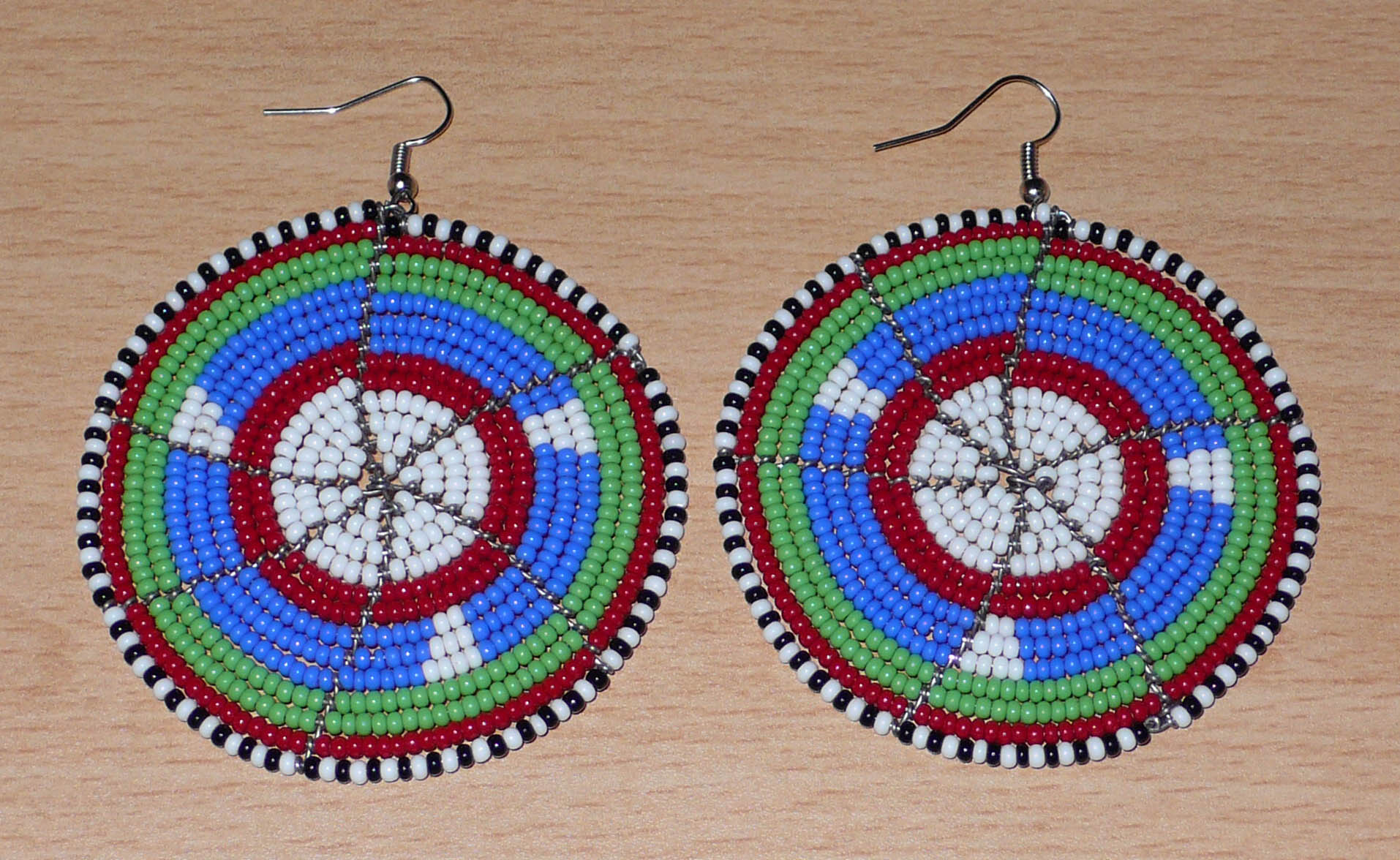 Boucles d'oreilles africaines ethniques Massai rondes composées de perles de rocaille bleues, vertes, rouges, blanches et noires. Elles mesurent 8 cm de long et 6,5 cm de large, et se portent sur des oreilles percées.