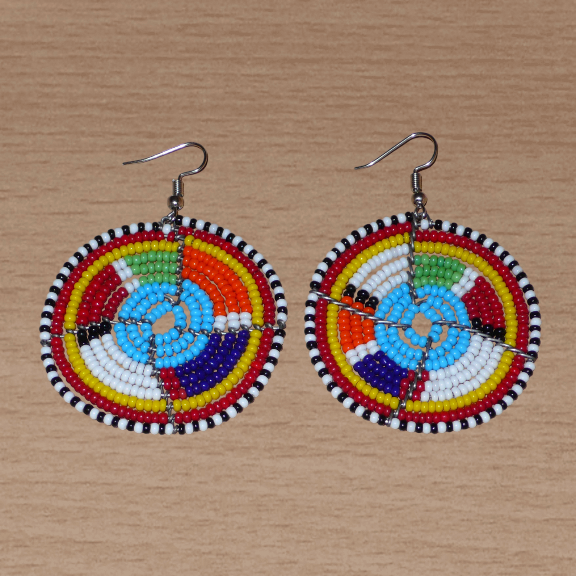 Boucles d'oreilles africaines bleues et multicolores à motifs ethniques traditionnels Massai, composées d'un tissage de perles de rocaille glissées sur des structures circulaires en fils de fer. De taille moyenne, ces boucles d'oreilles perlées mesurent 6.5 cm de long et 4.5 cm de large et se portent avec des crochets en acier inoxydable sur des oreilles percées.
