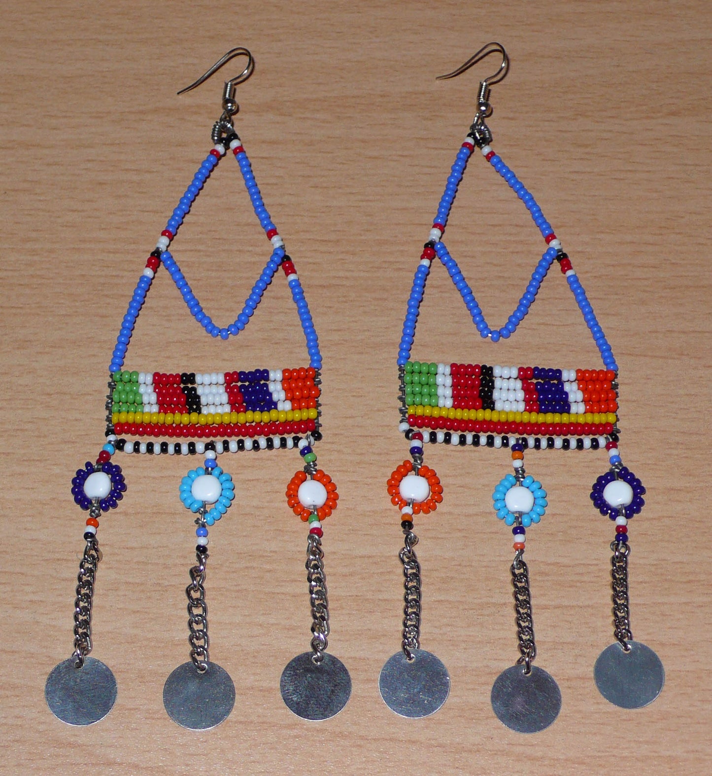 Boucles d'oreilles africaines Massai composées de perles de rocaille bleues et multicolores et de perles en plastique enfilées sur des structures à formes géométriques en fils de fer, et se prolongent de chaînettes décorées de disques métalliques. Surdimensionnées, elles mesurent 14.5 cm de long et se portent sur des oreilles percées.