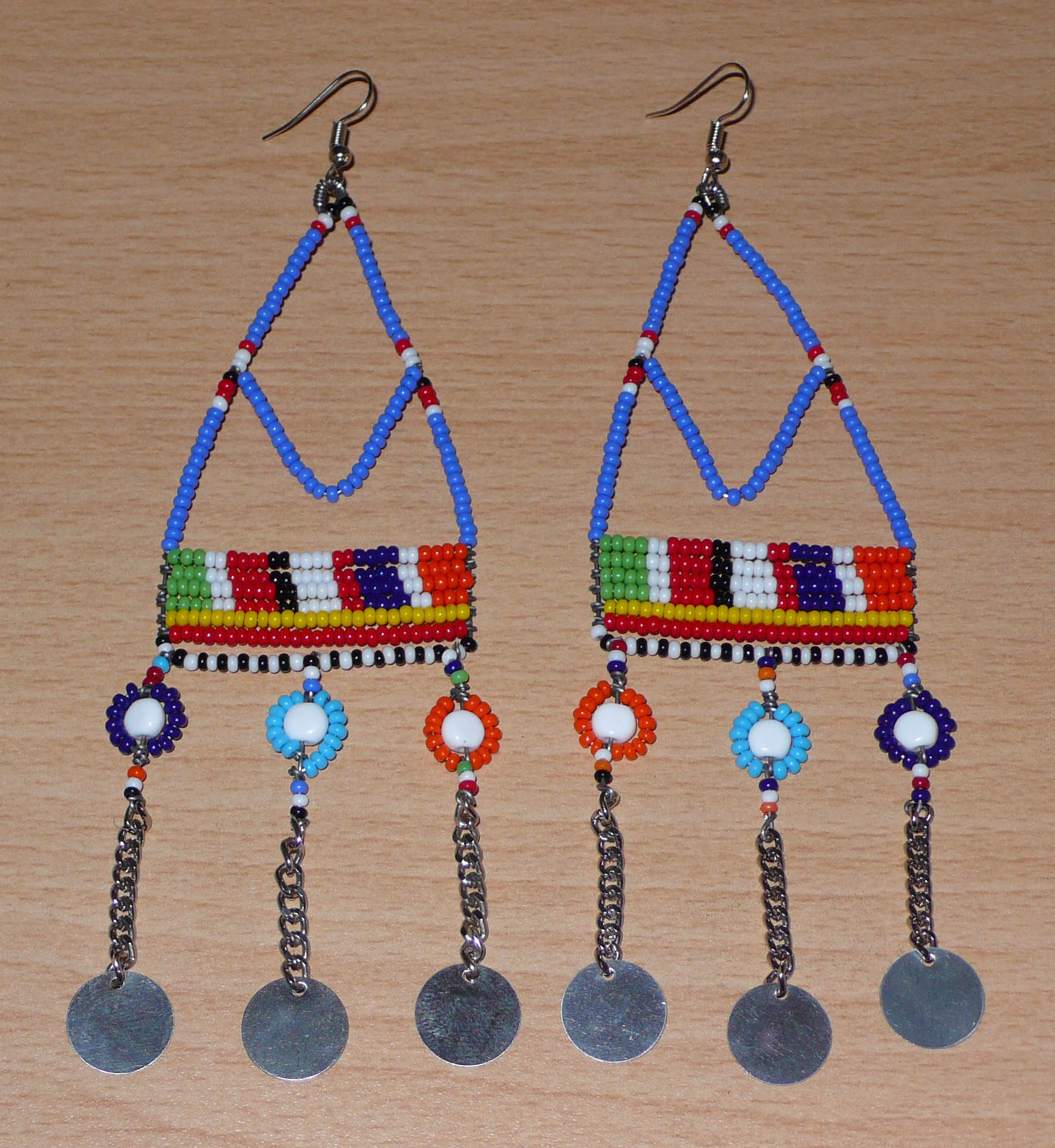 Boucles d'oreilles africaines Massai composées de perles de rocaille bleues et multicolores et de perles en plastique enfilées sur des structures à formes géométriques en fils de fer, et se prolongent de chaînettes décorées de disques métalliques. Surdimensionnées, elles mesurent 14.5 cm de long et se portent sur des oreilles percées.