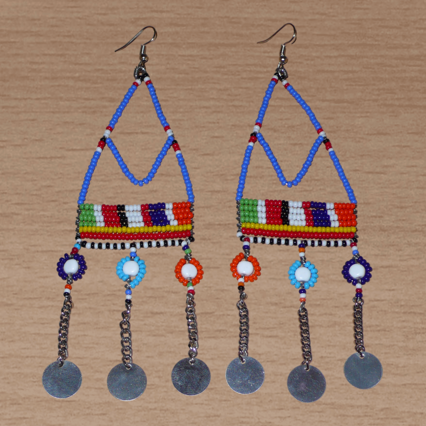 Boucles d'oreilles africaines Massai composées de perles de rocaille bleues et multicolores et de perles en plastique enfilées sur des structures à formes géométriques en fils de fer, et se prolongent de chaînettes décorées de disques métalliques. Surdimensionnées, elles mesurent 14.5 cm de long et se portent sur des oreilles percées.