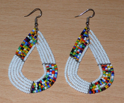 Boucles d'oreilles africaines multi-rangs blanches et multicolores aux accents évoquant le style bohème des années 70. Fabriquées au Kenya, elles se composent de perles de rocaille blanches et d'un mélange de perles multicolores glissées sur cinq rangées de fils de fer en forme de gouttes légèrement triangulaires. Ces boucles d'oreilles mesurent 8 cm de long et 4 cm de large ; elles se portent avec des crochets en acier inoxydable sur des oreilles percées.