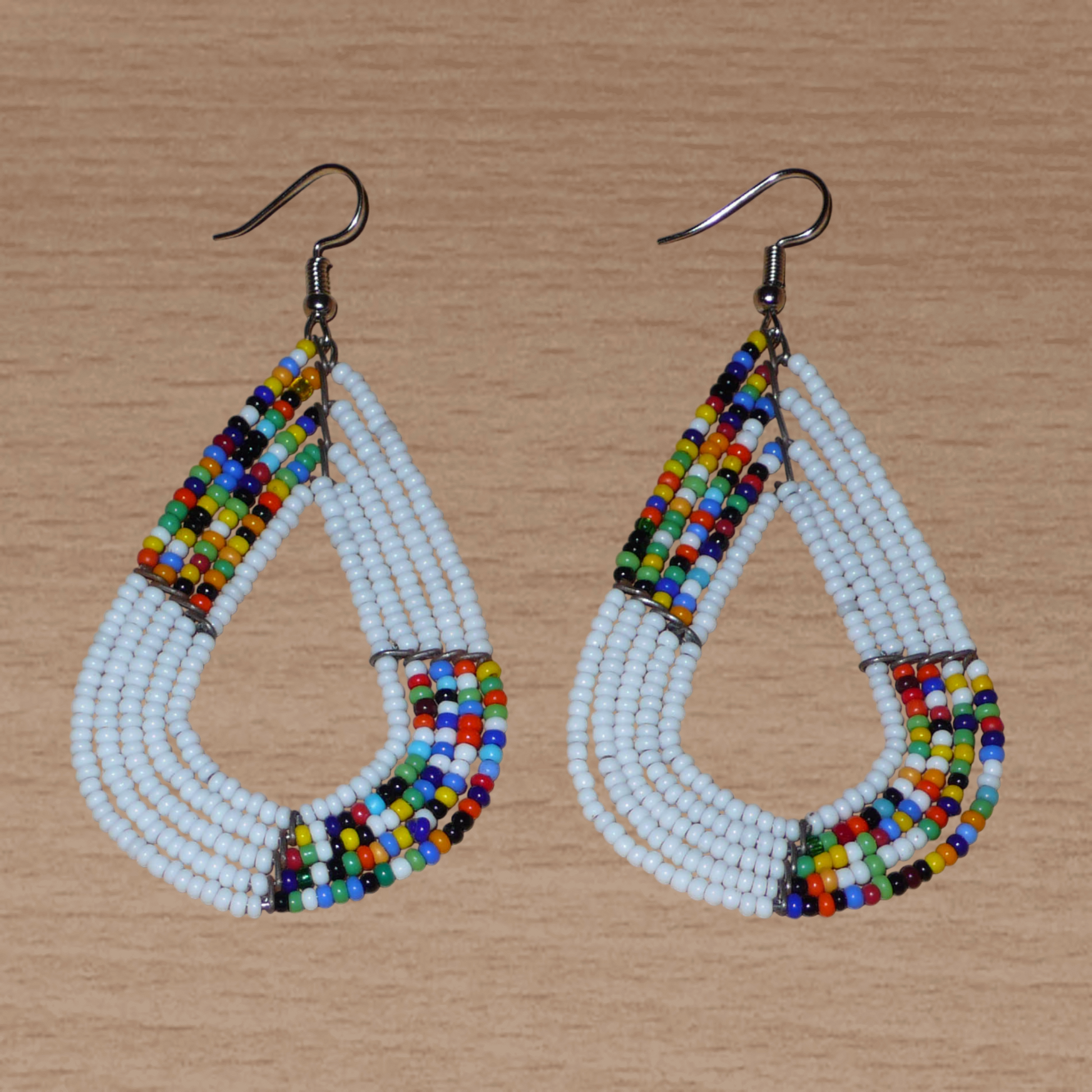 Boucles d'oreilles africaines multi-rangs blanches et multicolores aux accents évoquant le style bohème des années 70. Fabriquées au Kenya, elles se composent de perles de rocaille blanches et d'un mélange de perles multicolores glissées sur cinq rangées de fils de fer en forme de gouttes légèrement triangulaires. Ces boucles d'oreilles mesurent 8 cm de long et 4 cm de large ; elles se portent avec des crochets en acier inoxydable sur des oreilles percées.