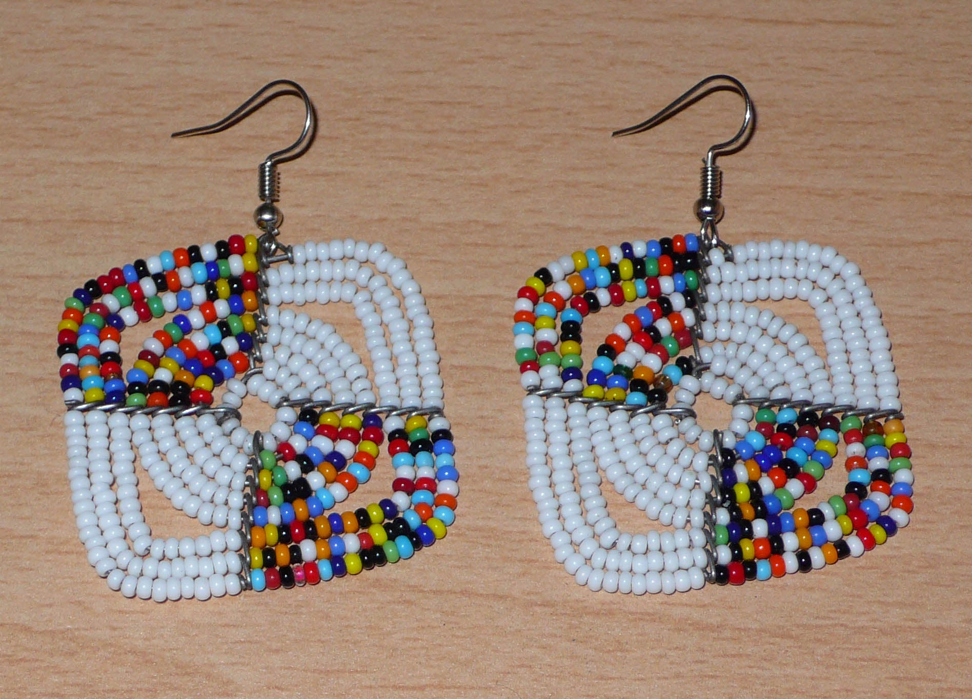 Boucles d'oreilles africaines Massai de confection artisanale kenyane composées de perles de rocaille blanches et d'un mélange de perles multicolores. Ces boucles d'oreilles aux accents exotiques mesurent 6.5 cm de long et 4.5 cm de large, et se portent avec des crochets en acier inoxydable sur des oreilles percées.
