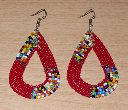 Boucles d'oreilles africaines Massai rouges et multicolores aux accents évoquant le style boho chic des années 70. Confectionnées au Kenya, elles se composent de perles de rocaille rouges et d'un mélange de perles multicolores glissées sur cinq rangées de fils de fer en forme de gouttes légèrement triangulaires. Ces boucles d'oreilles mesurent 8 cm de long et 4 cm de large ; elles se portent avec des crochets en acier inoxydable sur des oreilles percées.