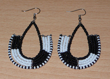 Boucles d'oreilles bicolores kenyanes en forme d'ancres, composées de perles de rocaille noires et blanches. Elles mesurent 7 cm de long et 4 cm de large, et se portent sur des oreilles percées.